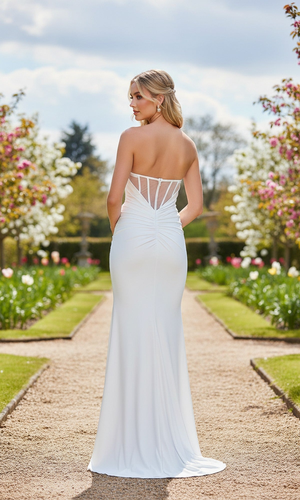 A woman stands on a garden path amid blooming flowers and trees, wearing the Strapless Ruched Tight Prom Gown: La Femme 33415 in white, its long, strapless design highlighting elegant ruched details as she faces away.